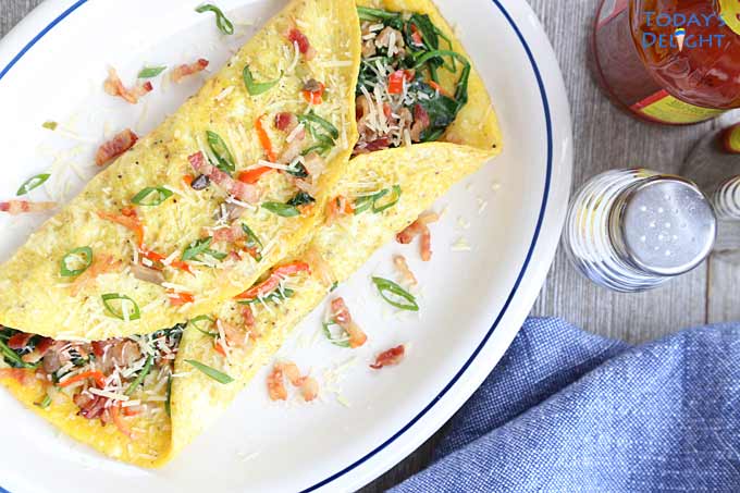 An omelette on a plate made with spinach, mushroom, red and green peppers and bacon wrapped in egg. Hot sauce, salt and pepper on the side.