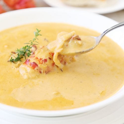 Lobster bisque soup with fresh thyme, a whole lobster tail and chunks of lobster on a spoon and a side of French bread.