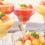 A big bowl of melon balls with cocktail sticks and 4 glasses of melon ball drink with mint.