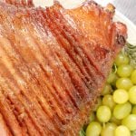 Hone baked spiral cut ham on a platter decorated on the side with fresh rosemary and green grapes.