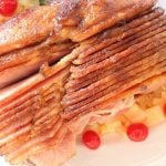 Pineapple honey glazed spiral cut ham on a platter with fresh rosemary, cherries and pineapple.
