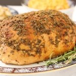 Seasoned fully cooked boneless turkey breast in a platter with fresh rosemary and baby carrots, turkey gravy and side dishes.