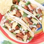 Three tacos al pastor on a plate with a side of tortilla chips and salsa.