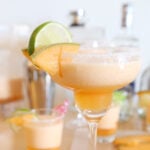 One cantaloupe margarita in a margarita glass and two are in cocktail glasses decorated with a couple slices of cantaloupes and lime. A shaker and bottles behind the glasses.