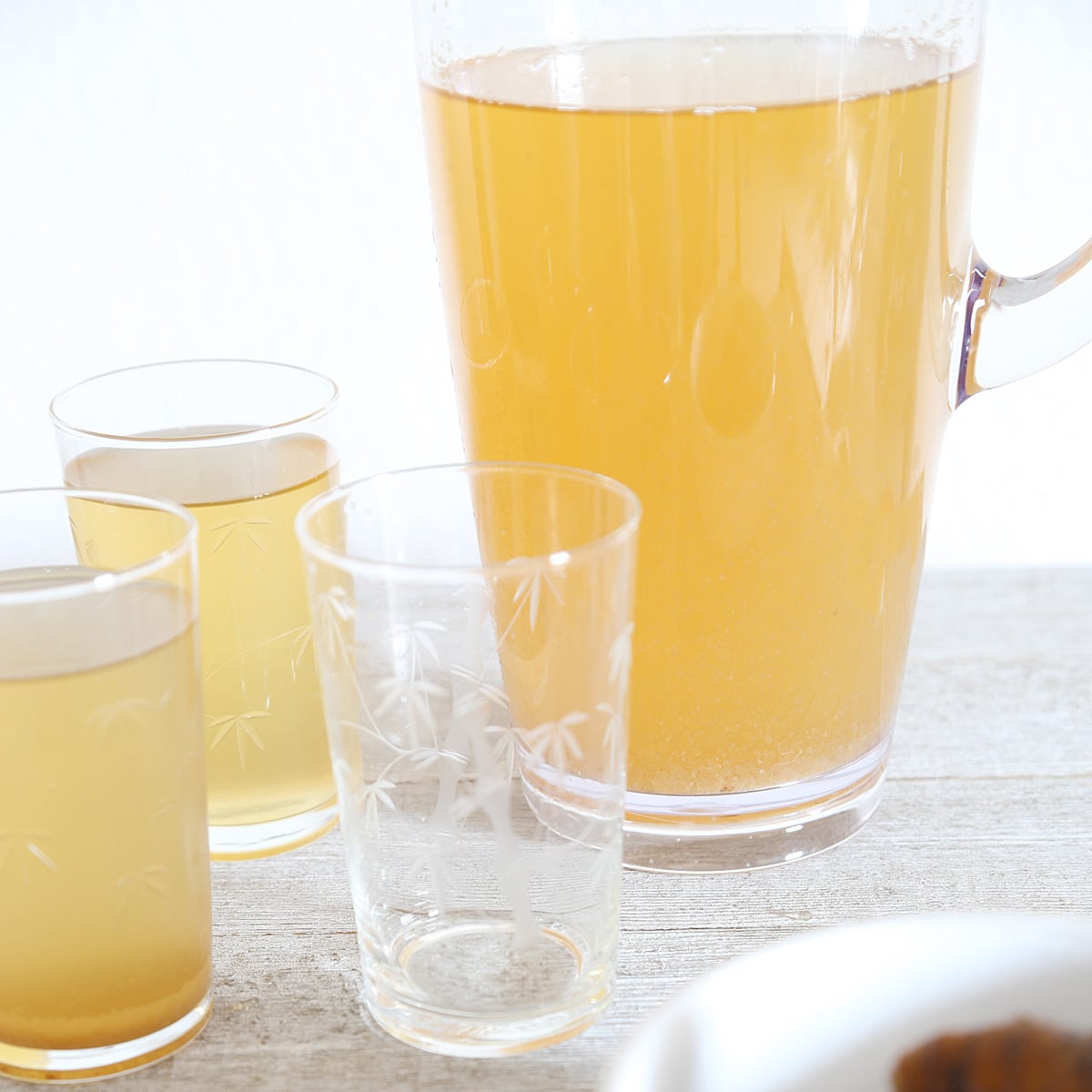 A pitcher of cold turmeric tea poured into 2 glasses and some whole unpeeled turmerics in a bowl.
