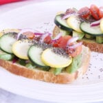 Two slices of toasted bread topped with cucumber, tomato, avocado, red onion, zucchini sprinkled with chia seeds on a plate.