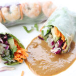 One piece vegetable and shrimp spring roll and another one cut in half laying on peanut sauce on a plate.