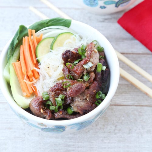 A bowl of Vietnamese grilled pork with rice noodles, thinly sliced carrots, cucumber, napa leaf, sprinkled with green onions. A chopstick, napkin, and another bowl nearby.