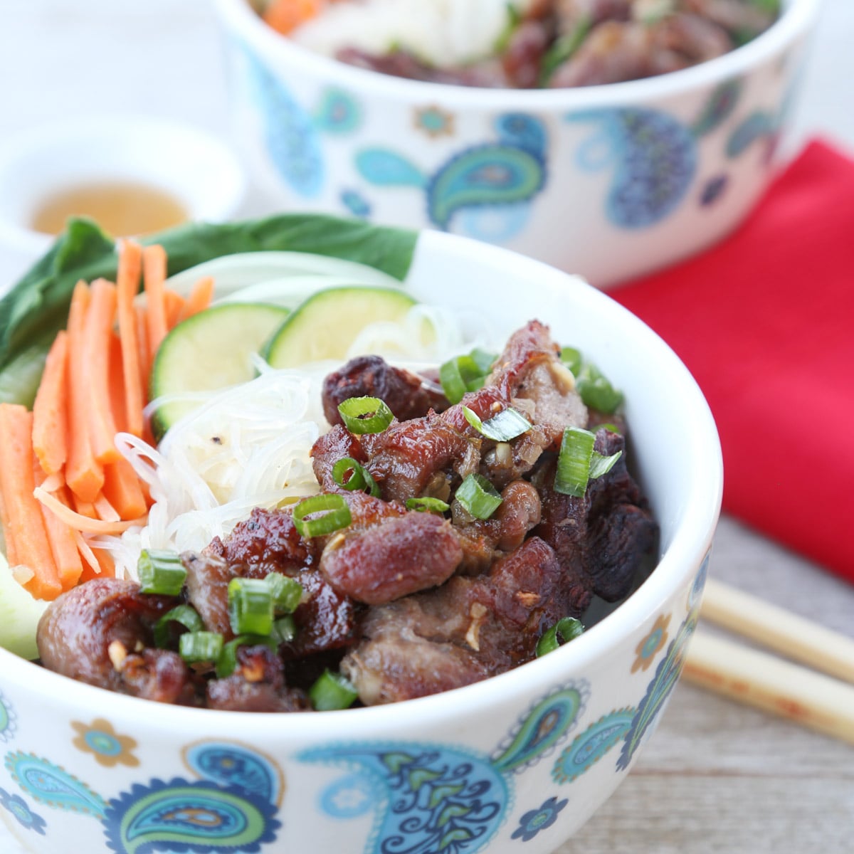 Two bowls of Vietnamese grilled pork with rice noodles, thinly sliced carrots, cucumber, napa leaf, sprinkled with green onions. A side of fish sauce, chopstick, and napkin nearby.