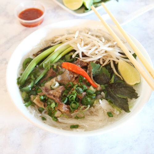 A big serving of Vietnamese beef pho consisting of beef broth, rice noodles, herbs, and meat, and garnished with bean sprouts, Thai basil, jalapeno, green onions, chili peppers, and wedge of lime. Chopstick on the bowl, a serving of sriracha and more garnishes on the side.