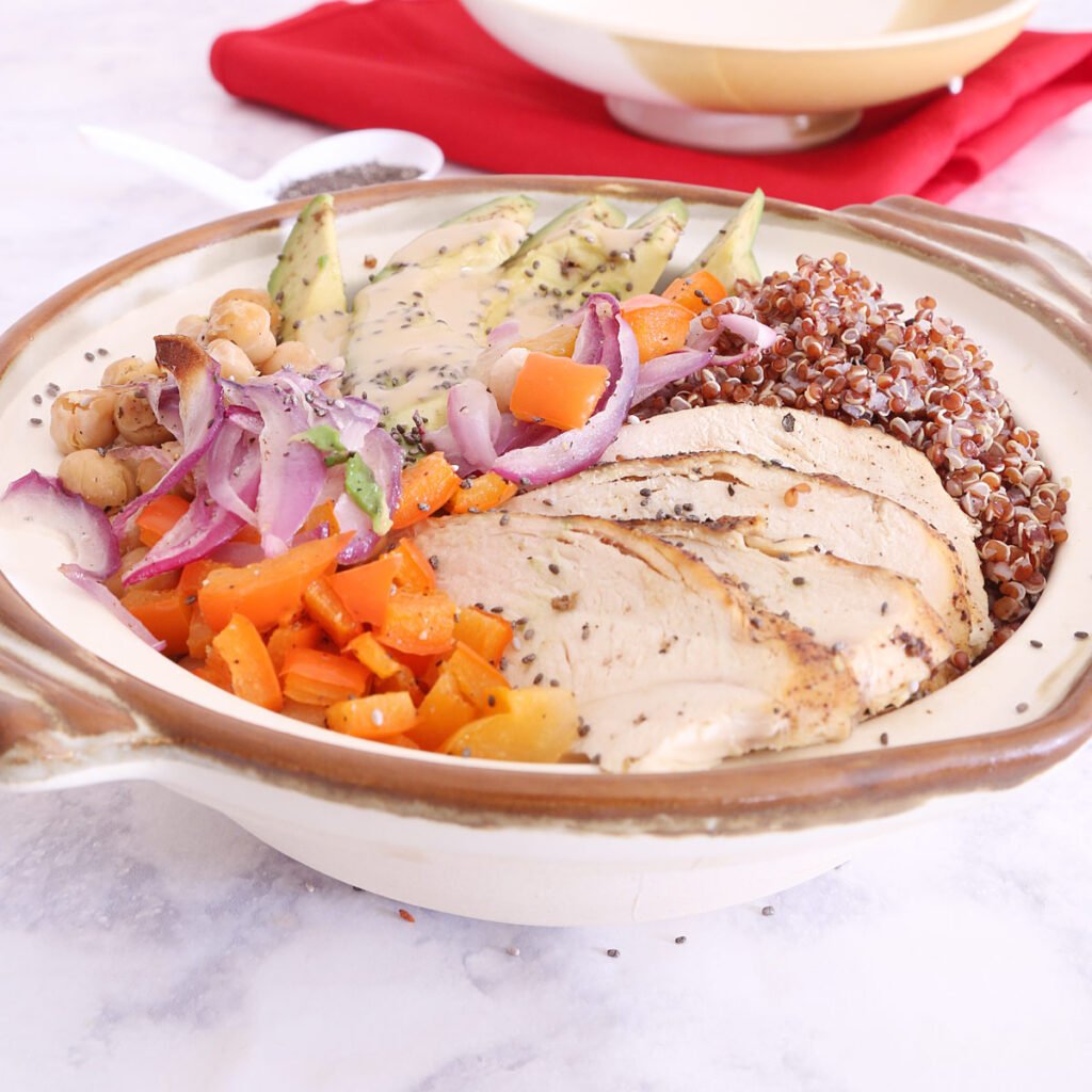 Chicken and Quinoa Bowl has roasted onion, red bell pepper, garbanzos, sliced avocado, lemon pepper chicken, chia seeds drizzled with creamy tahini dressing. A tablespoon of chia seeds and the bowl cover sits on a red napkin in the background.