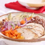 Chicken and Quinoa Bowl has roasted onion, red bell pepper, garbanzos, sliced avocado, lemon pepper chicken, chia seeds drizzled with creamy tahini dressing. The bowl cover sits on a red napkin in the background.