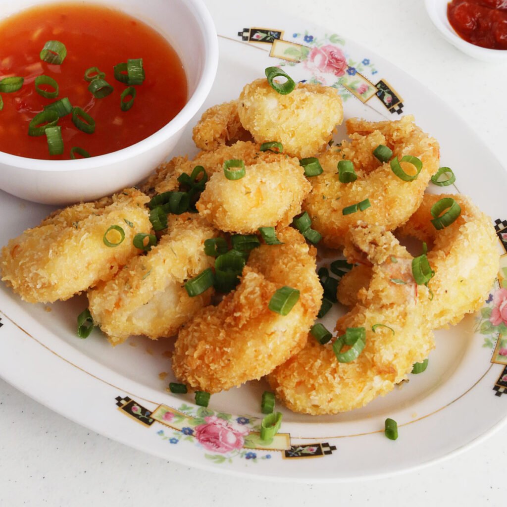 Eight crispy breaded shrimp on a platter with a side of sweet and sour sauce on a small bowl, and both are garnished with green onions. A ketchup on a saucer on the side.